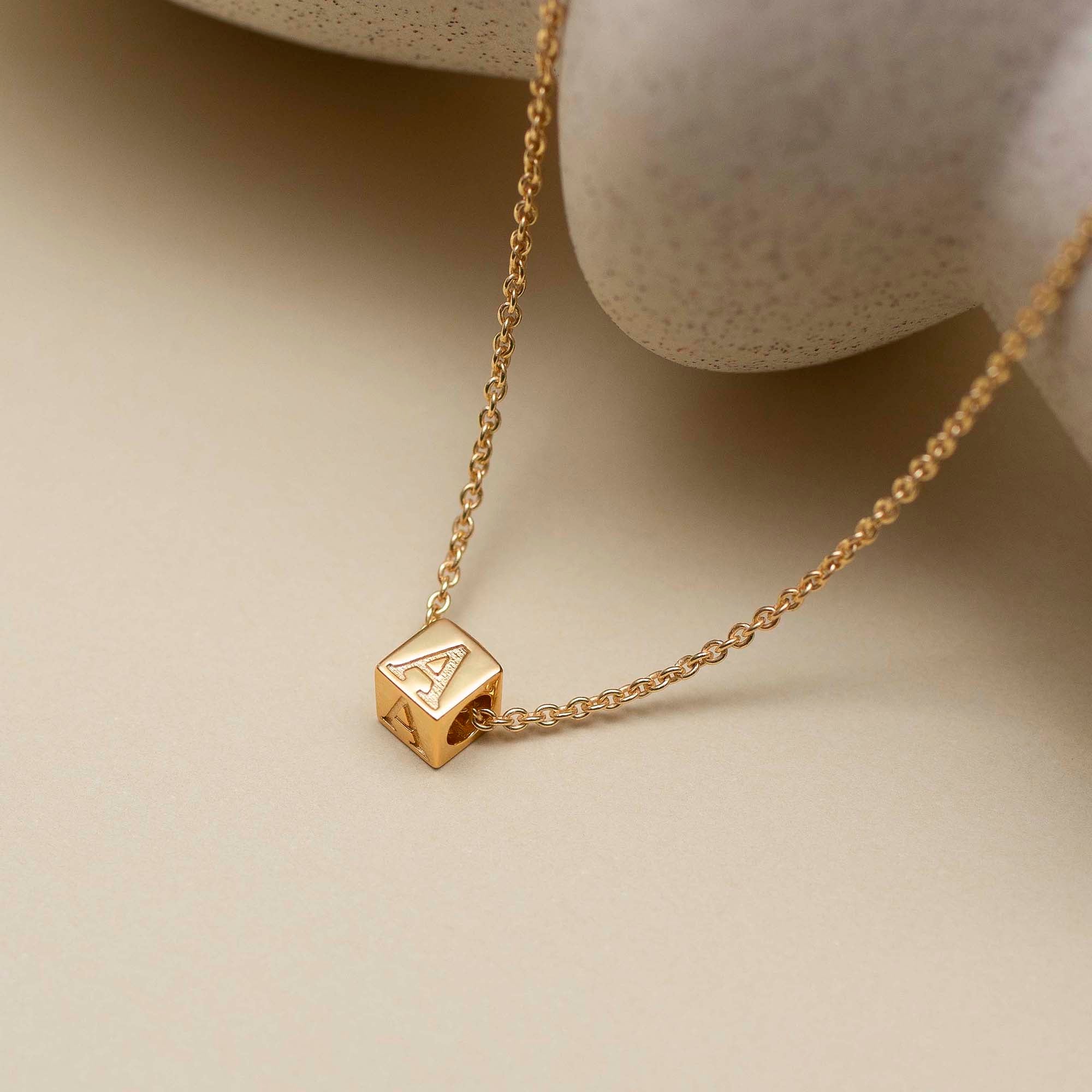 Gold necklace with a cube pendant featuring an 'A' on a beige background
