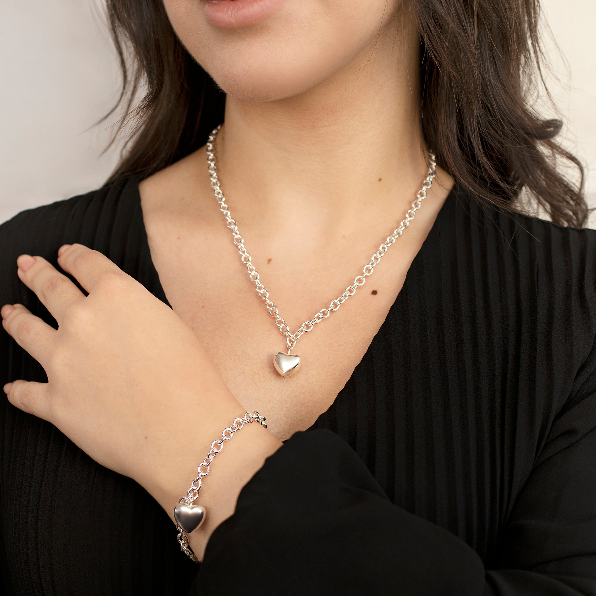 Person wearing a silver necklace and bracelet with heart pendants on a plain background