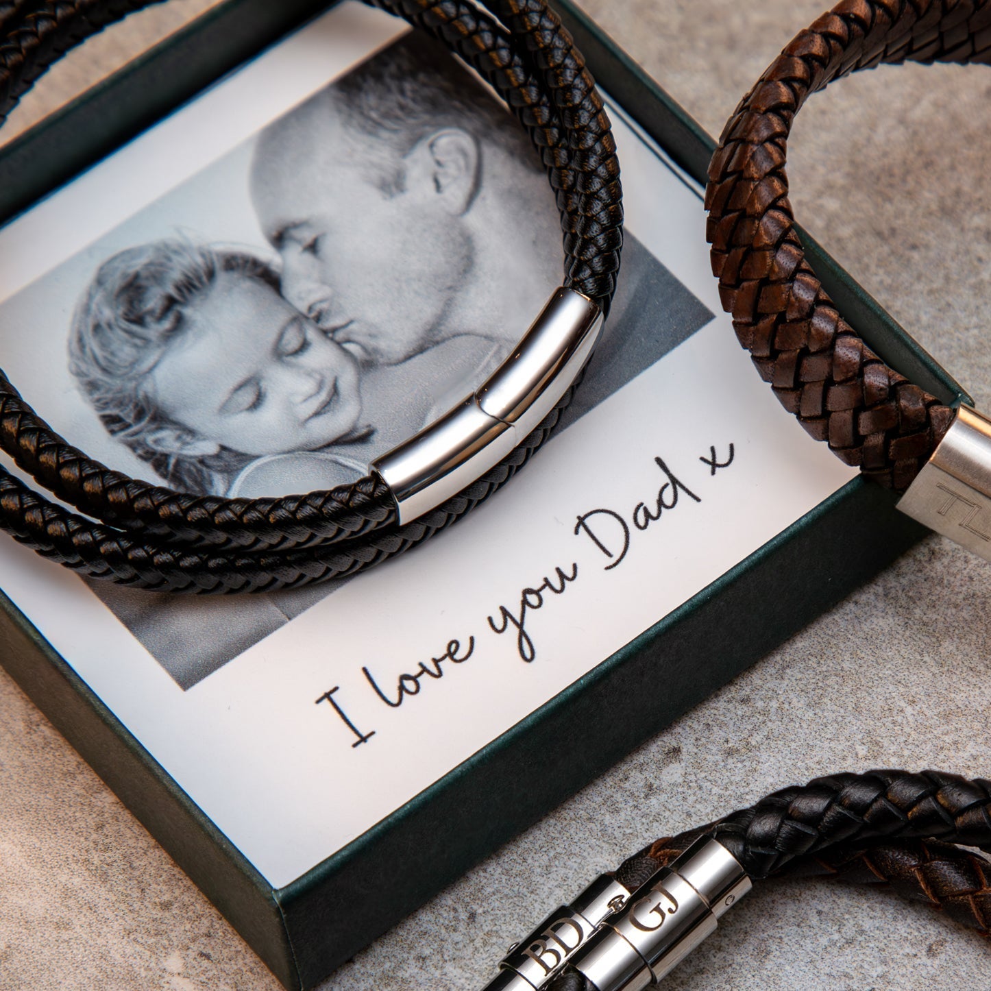 Two braided leather bracelets with silver clasps on a textured surface next to a photo frame with 'I love you Dad' text.