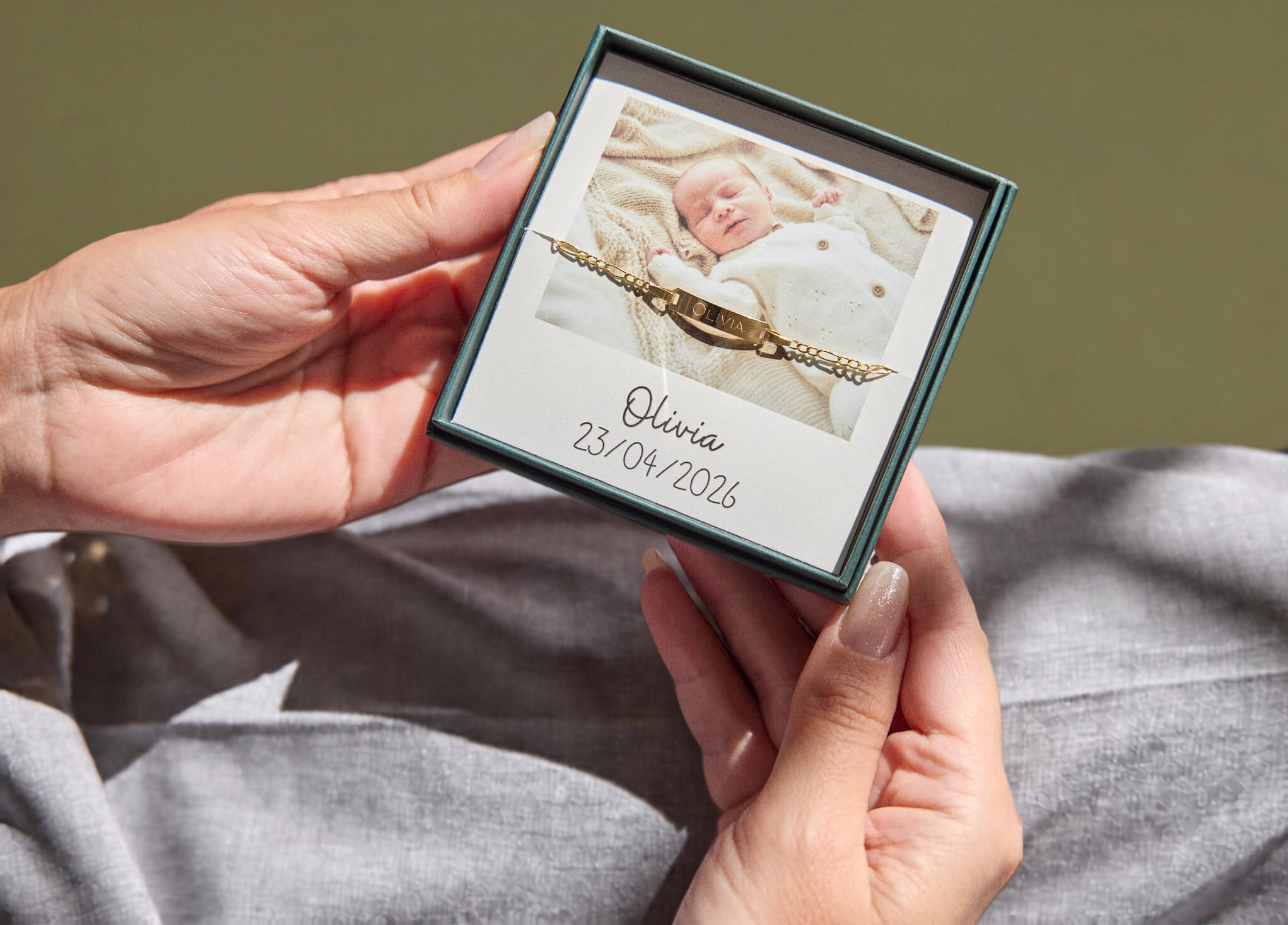 A person holding a small square gift box containing a personalized baby bracelet. The box has a dark teal border and a white insert showing a photo of a newborn baby and the name 'Olivia' with the date '23/04/2026'.